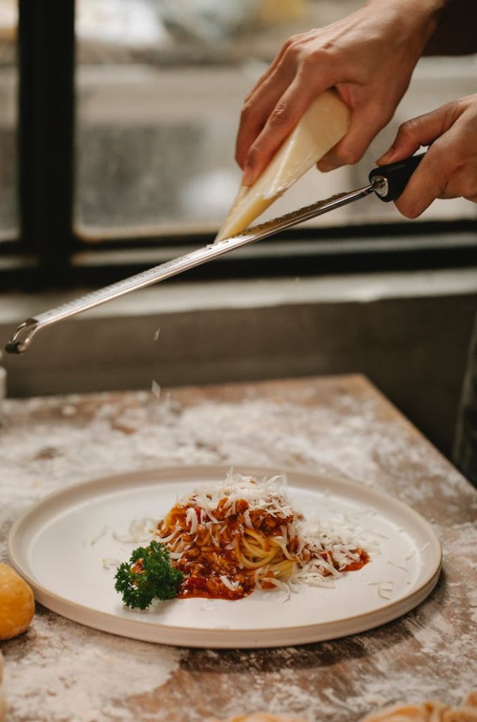 Crop anonymous chef grating cheese on freshly cooked spaghetti with tomato sauce and parsley served on table in kitchen
